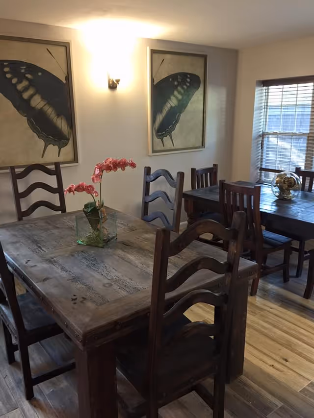 Interior view of a dining room with two wooden tables and several matching wooden chairs. One table has a glass vase with pink flowers, and the other table has a decorative globe. Two large butterfly paintings hang on the wall, and a window with blinds lets in natural light.