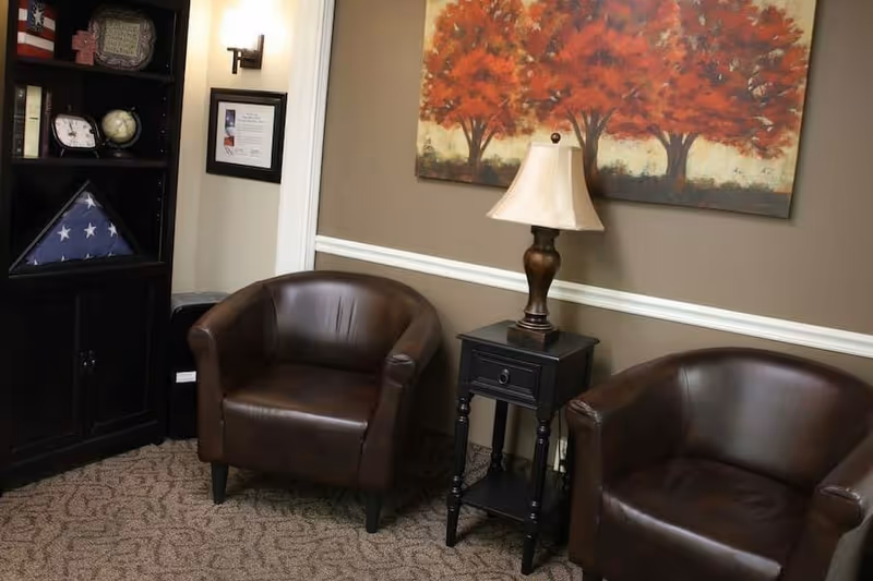 A cozy sitting area with two brown leather armchairs separated by a small black side table with a lamp. Behind the table is a painting of three red-leaved trees on a beige wall with white trim. To the left, there is a dark wooden bookshelf with various items including a folded American flag in a triangular display case, a clock, and a globe.