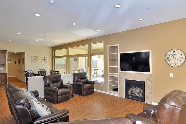 Spacious common living room with leather recliners, a wall-mounted TV above a fireplace, large windows, and a dining area visible in the background.