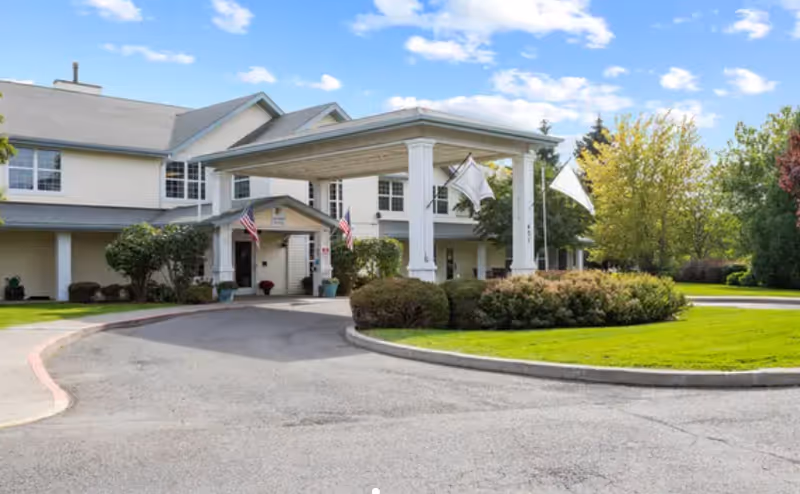 Exterior view of Sullivan Park Senior Living facility showing a two-story building with a covered entrance, surrounded by well-maintained landscaping including bushes and trees under a partly cloudy sky.