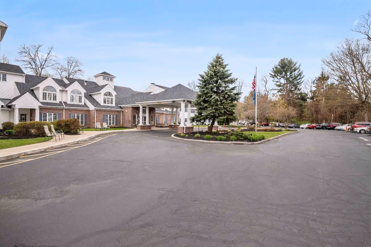 Exterior view of Middlebrook Farms at Trumbull showing a large paved driveway and parking area with several parked cars. The building has a brick and white siding facade with multiple windows and a covered entrance. There are landscaped areas with bushes and a large evergreen tree near the entrance, and an American flag is visible on a flagpole.