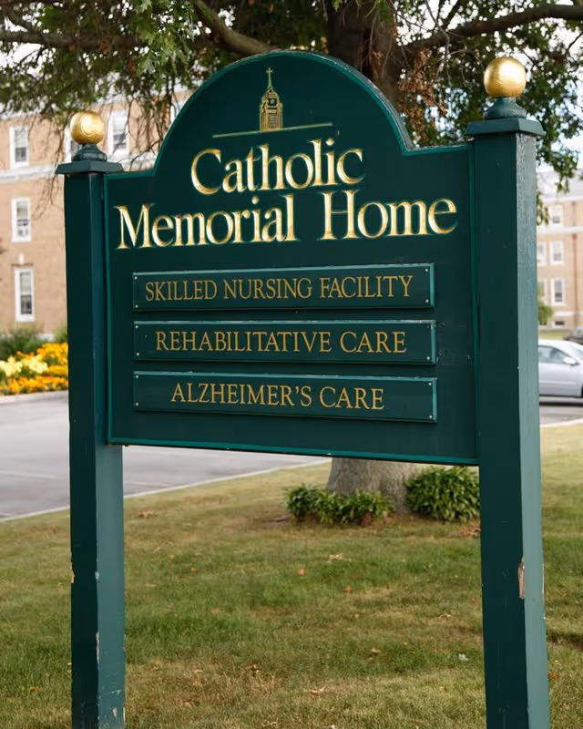 Green roadside sign reading 'Catholic Memorial Home' listing Skilled Nursing Facility, Rehabilitative Care, and Alzheimer's Care in front of a lawn and building.
