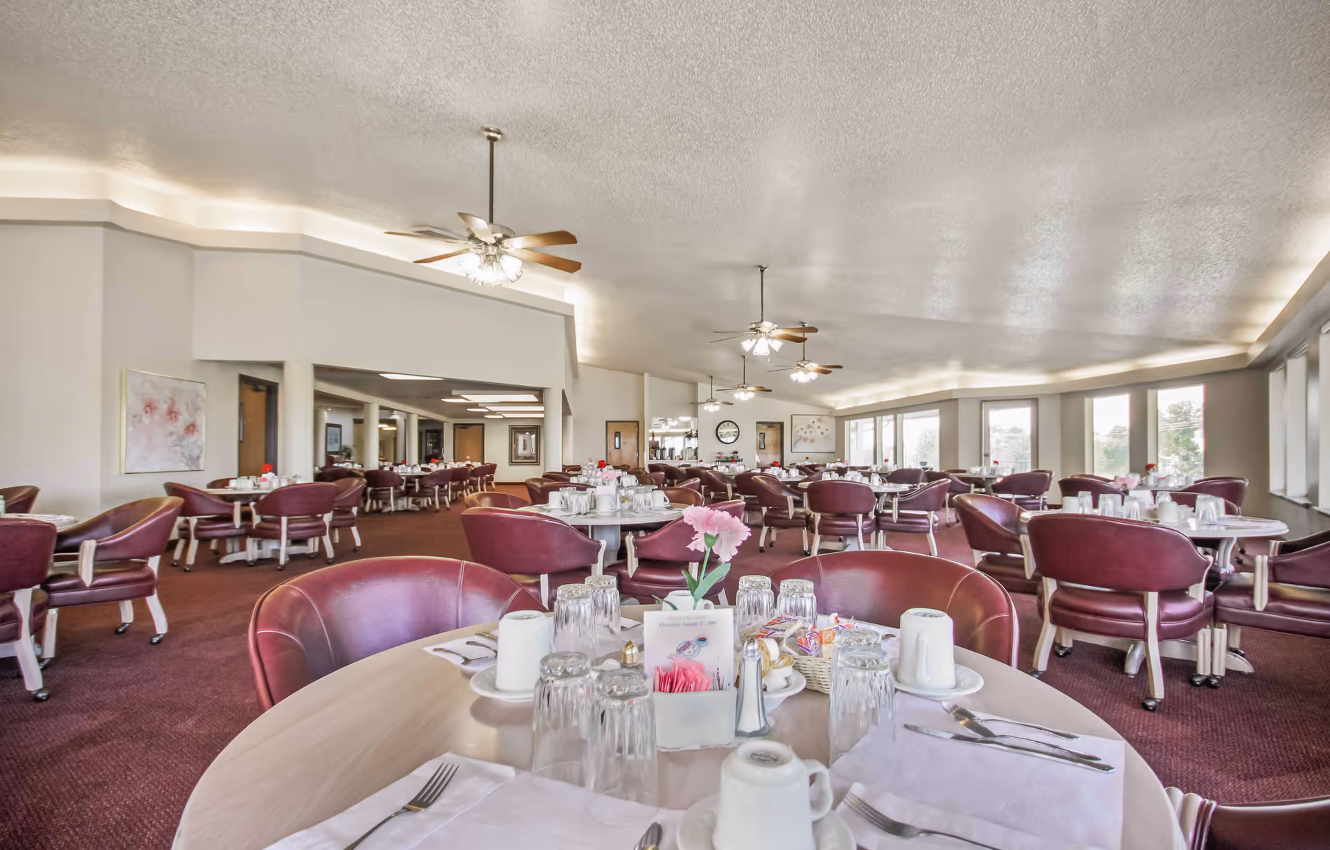 A spacious dining room with multiple round tables set with white cups, glasses, silverware, and napkins. Each table has a small centerpiece with pink flowers. The room features maroon chairs, ceiling fans with lights, large windows letting in natural light, and artwork on the walls.