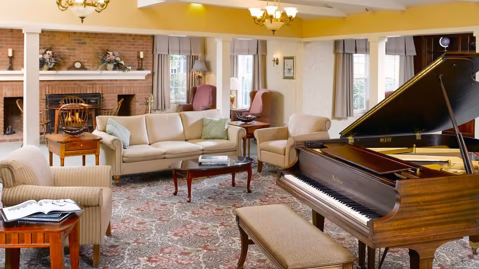 A cozy senior living facility common area featuring a grand piano with a cushioned bench, several upholstered armchairs, a beige sofa with light green pillows, a wooden coffee table, and a brick fireplace decorated with floral arrangements and candles. The room has large windows with curtains, warm lighting from ceiling fixtures, and a patterned carpet.