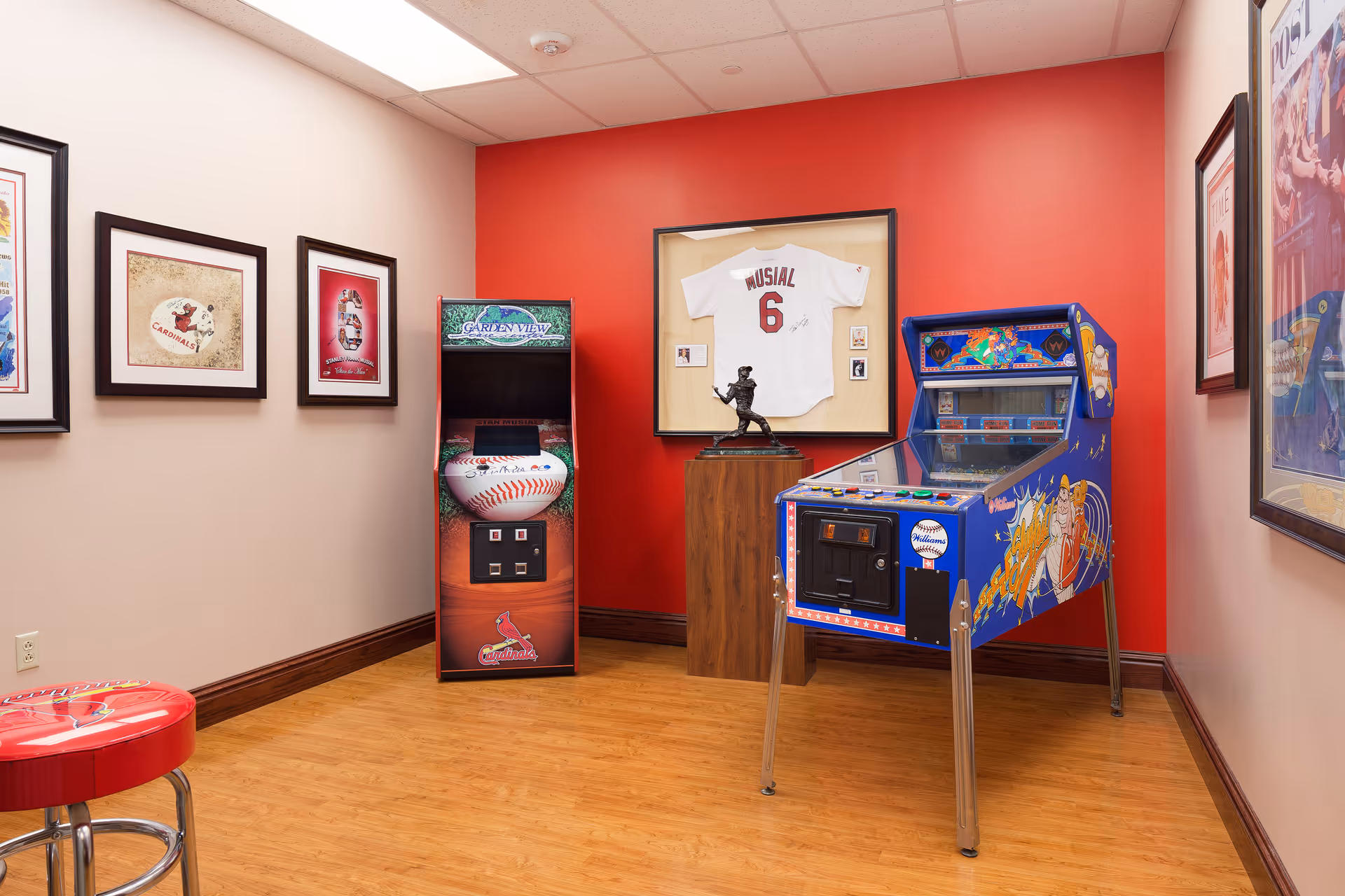 A small game room with a wooden floor and red and beige walls. The room features a blue pinball machine, a baseball-themed arcade game, a framed white baseball jersey with the name Musial and number 6, a small statue on a wooden pedestal, and several framed pictures on the walls. A red stool with a Cardinals logo is partially visible in the foreground.