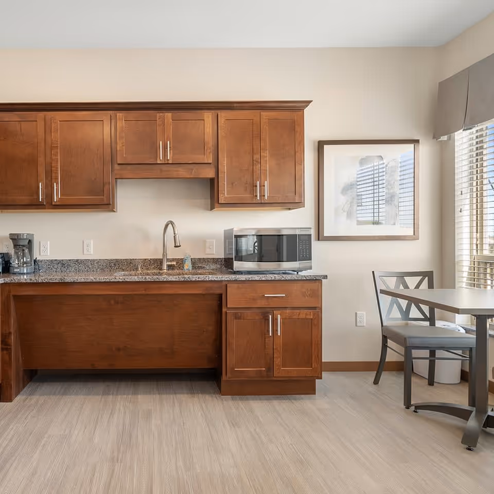 A kitchen area with wooden cabinets, a granite countertop, a stainless steel sink with a faucet, a coffee maker, and a microwave. To the right, there is a small dining table with a chair next to a window with blinds and a framed picture on the wall.