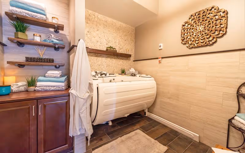A well-lit bathroom featuring a large white walk-in bathtub with multiple jets, a beige tiled wall with a decorative wooden shelf holding small plants and towels, a wooden cabinet with neatly folded towels and decorative items, a white bathrobe hanging on a hook, and a decorative golden wall art piece above the bathtub.