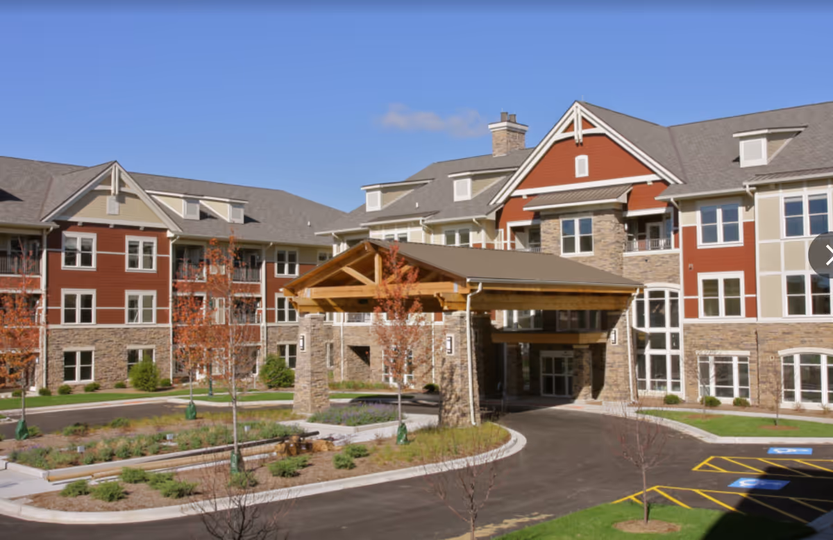 Exterior view of a large senior living facility building with stone and red siding, multiple windows, and a covered entrance with wooden beams. The driveway curves around landscaped areas with young trees and shrubs under a clear blue sky.