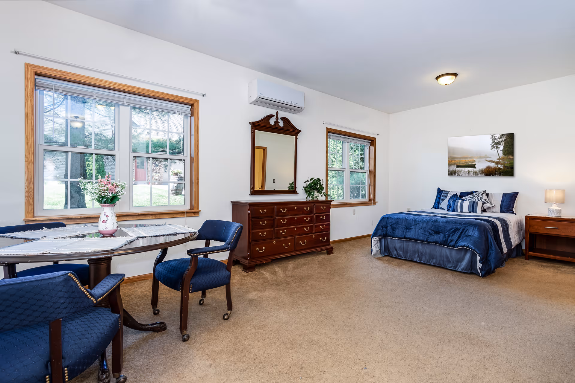 Bright bedroom in a senior living suite featuring a bed, dresser with mirror, and two windows.