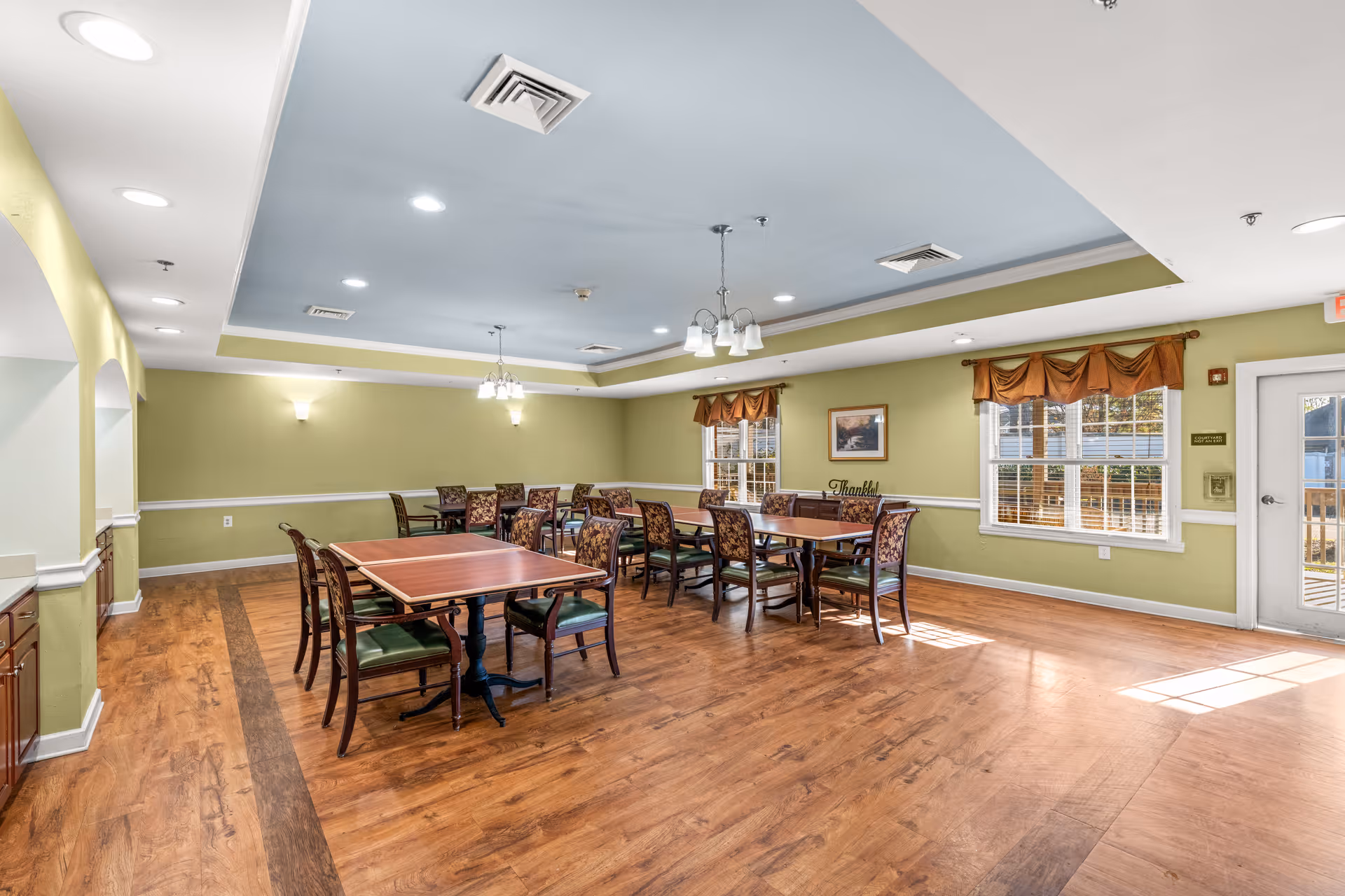A bright dining room with wooden floors, green walls, and a light blue ceiling. The room contains several wooden tables with floral upholstered chairs arranged neatly. Two windows with orange valances let in natural light, and there are ceiling lights and wall sconces providing additional illumination. A door with a window is visible on the right side.