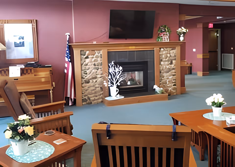 A cozy assisted living common area with wooden chairs and tables, a stone and wood fireplace with a TV mounted above it, an American flag in the corner, and floral decorations on the tables and mantel. The walls are painted a warm reddish color and the carpet is blue.