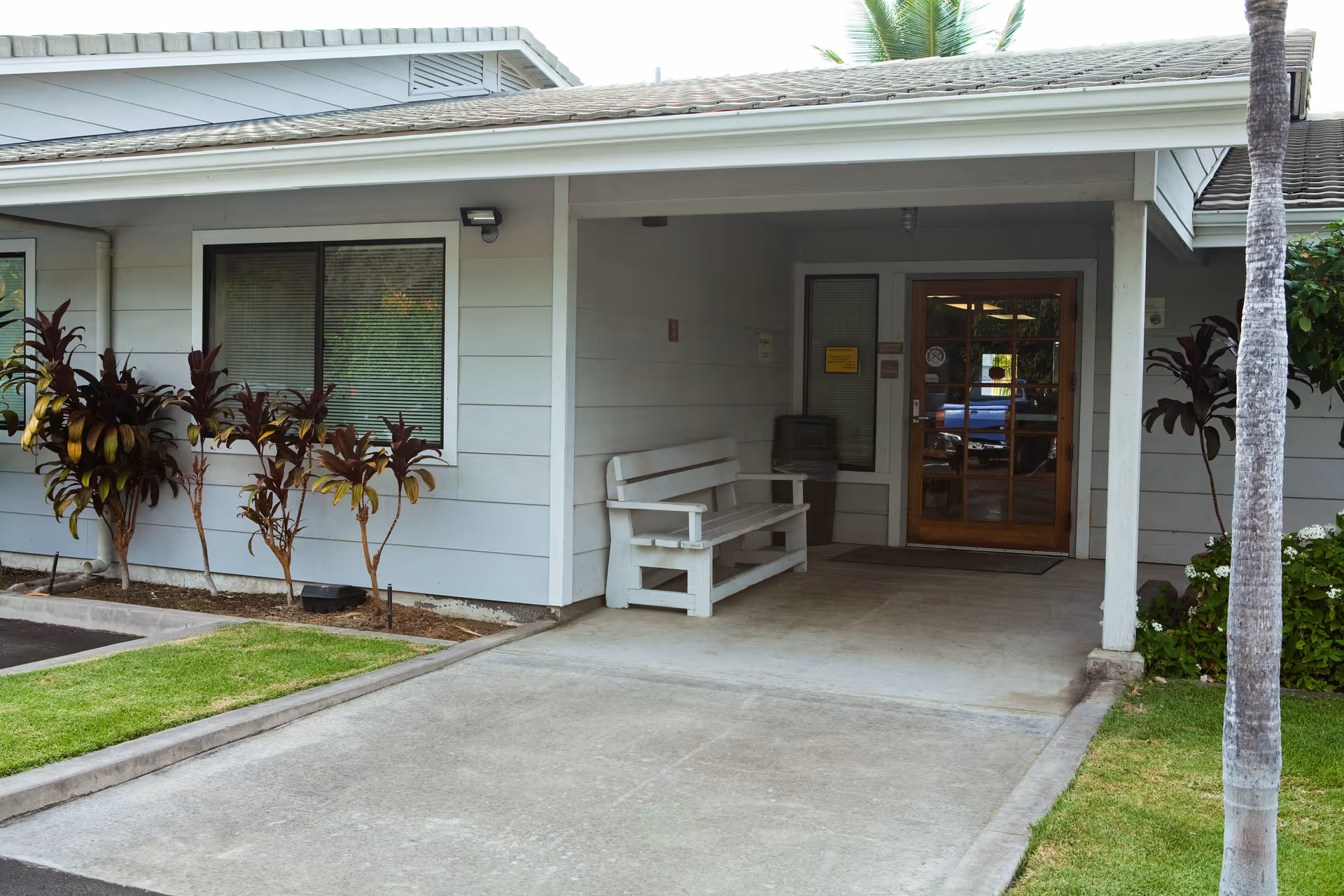 Covered entrance of a single-story building with a white bench, glass door, and landscaping.