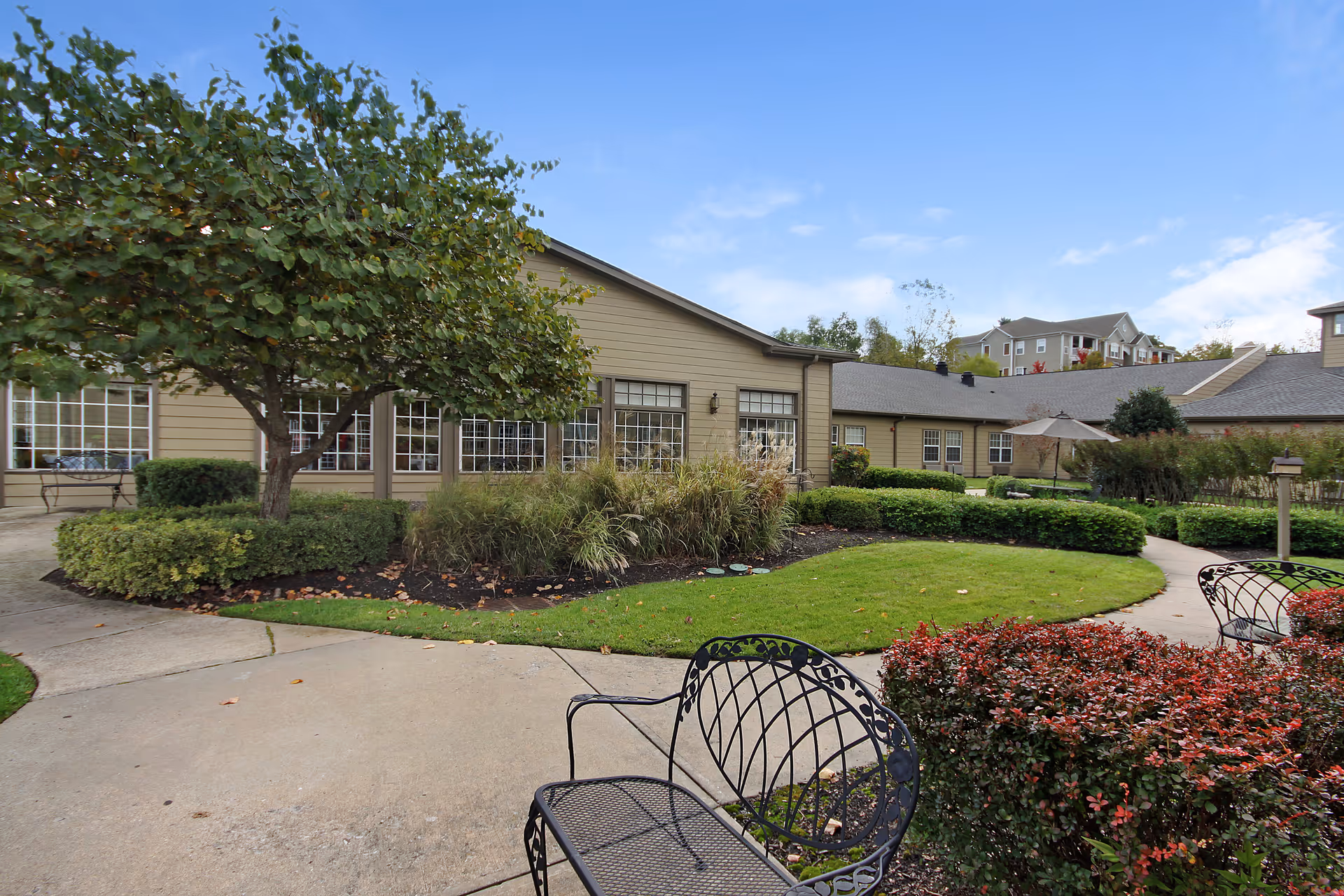 A landscaped courtyard with metal chairs, paved walkways, trimmed hedges and a single-story building under a blue sky.