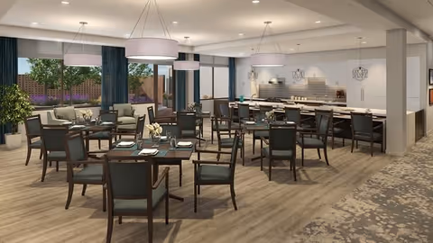 A spacious dining room in a senior living facility with multiple wooden tables and chairs arranged neatly. The room features large windows with dark curtains, modern pendant lights hanging from the ceiling, and a kitchen area with white cabinetry and a tiled backsplash in the background. The floor is covered with light wood and a patterned carpet near the entrance.