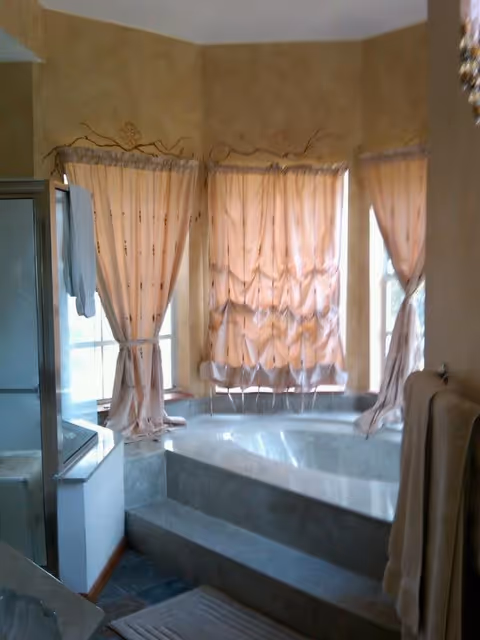 Bathroom with a sunken tiled bathtub accessed by steps beneath three bay windows covered with peach curtains, a glass shower on the left and towels hanging on the right.