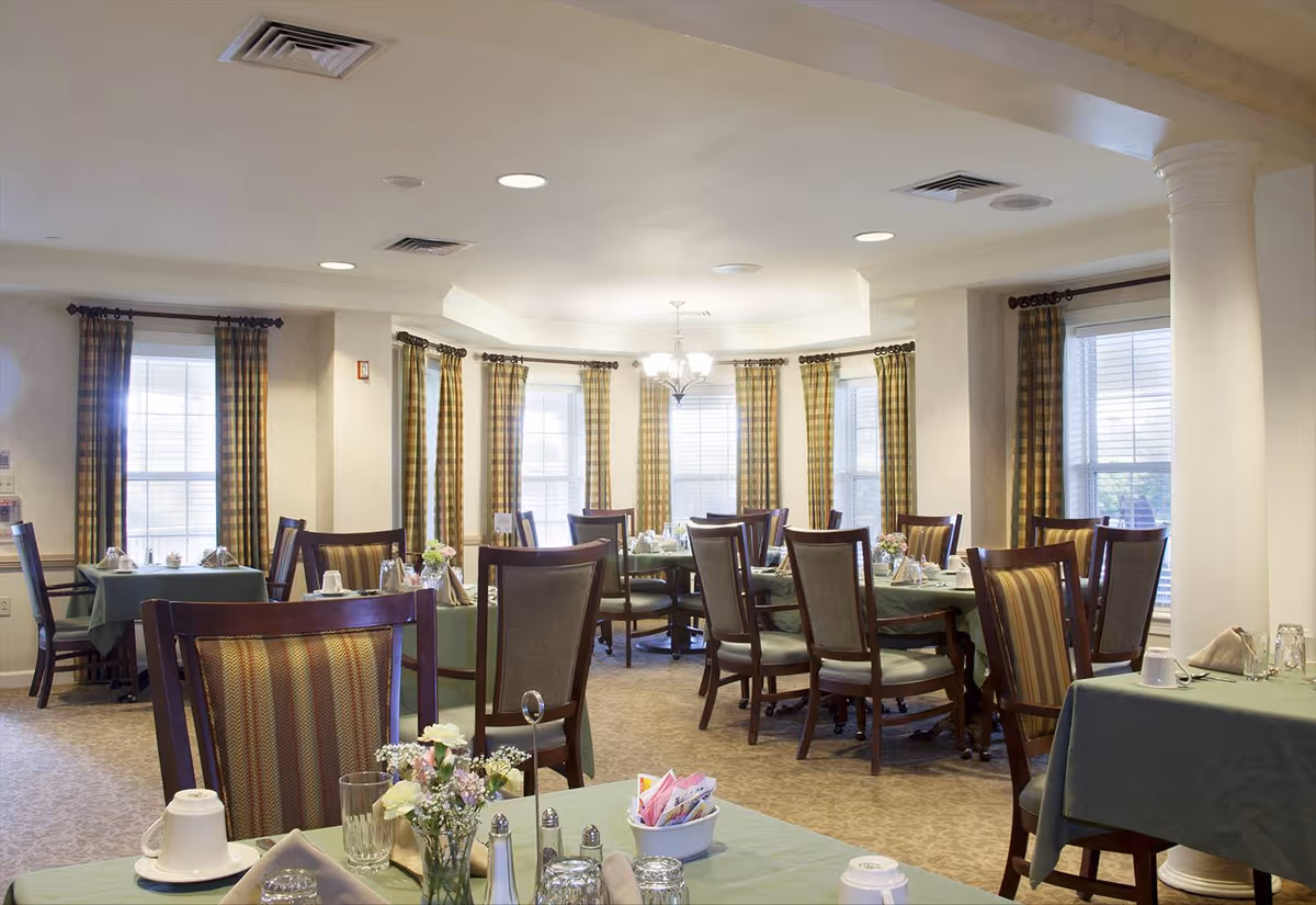 A bright dining room with multiple tables covered in green tablecloths, each set with cups, glasses, napkins, and small flower arrangements. The room has large windows with plaid curtains and a chandelier hanging from the ceiling.
