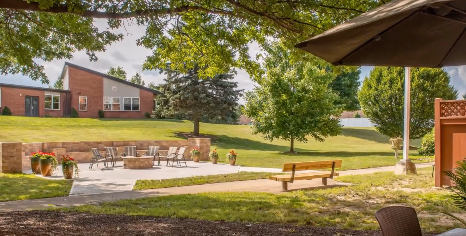 Outdoor patio area with several chairs arranged around a circular fire pit, surrounded by a low brick wall. There are potted plants with red flowers placed around the patio. In the background, there is a grassy hill with trees and a brick building. A wooden bench is positioned along a paved walkway, and a large umbrella partially shades the foreground.