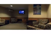 A dimly lit senior living common room with upholstered armchairs, a wall-mounted TV, and framed artwork on the wall.