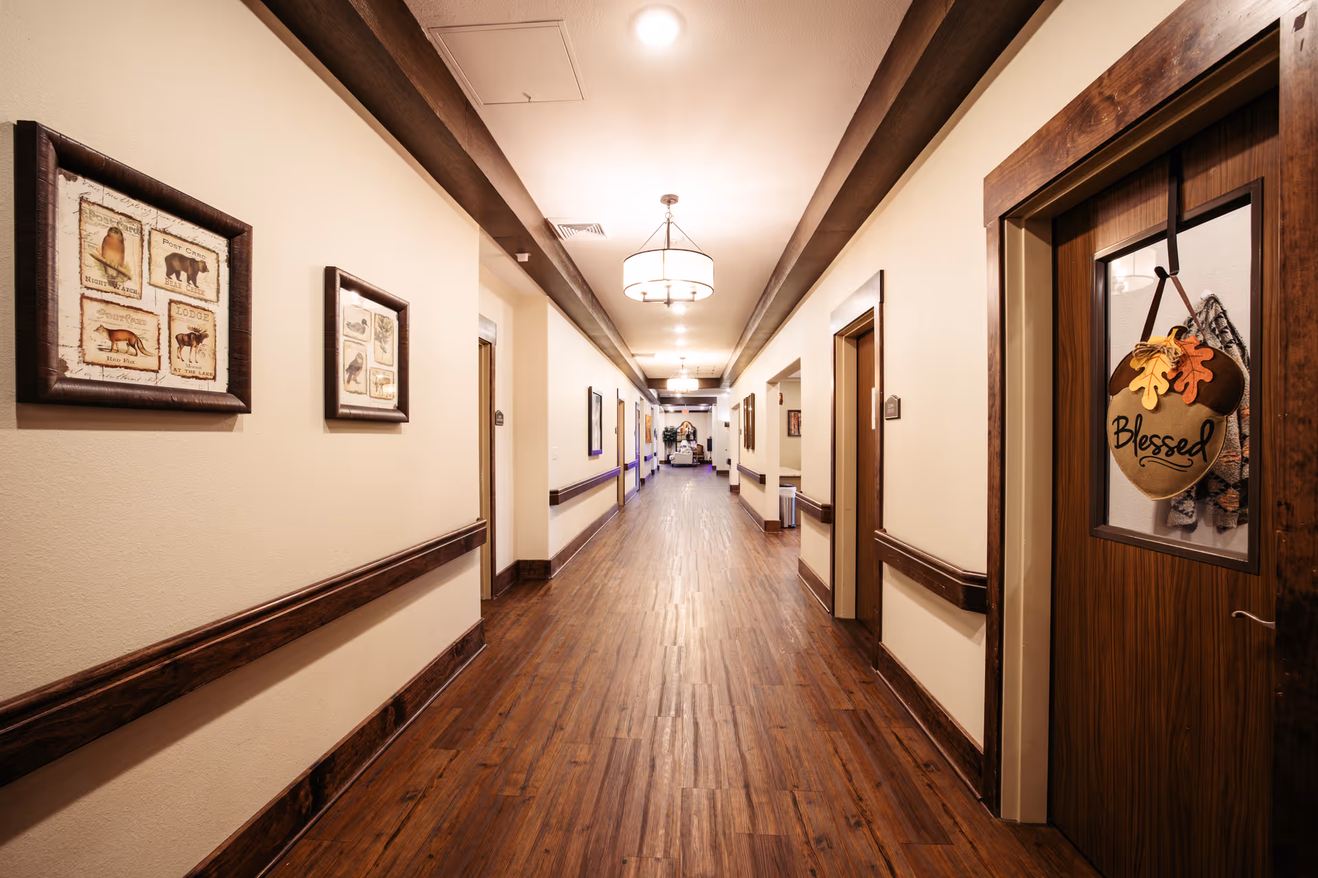 A long, well-lit hallway in a nursing and rehabilitation center with wooden floors and beige walls. The hallway features wooden handrails on both sides and framed pictures on the walls. There are several doors along the hallway, one of which has a decorative sign that reads 'Blessed' hanging on it. Ceiling lights provide warm illumination.