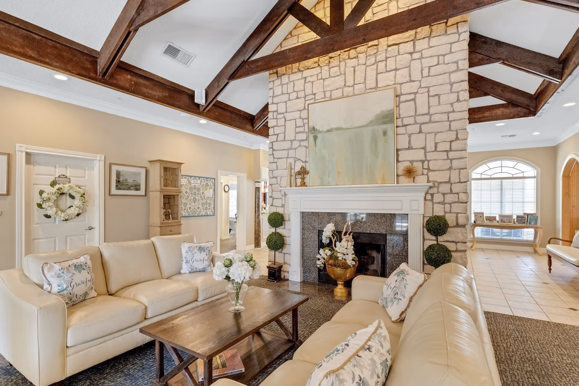 Spacious living room with cream leather sofas, a stone fireplace topped by artwork, exposed wooden ceiling beams, and a central coffee table.