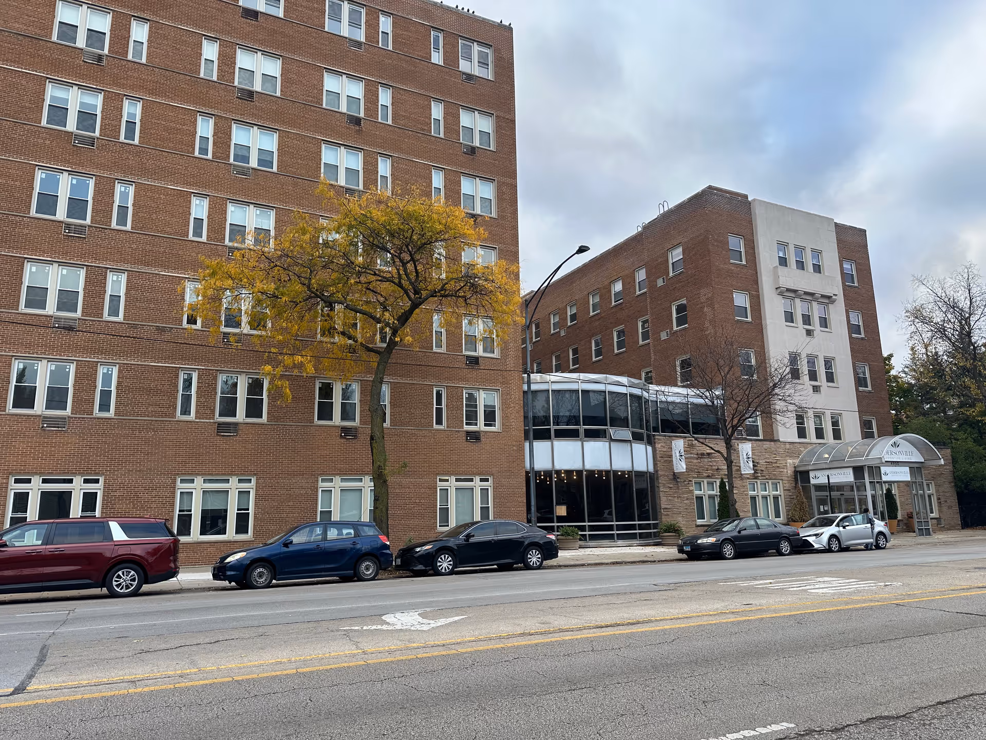 Exterior view of a multi-story brick building with numerous windows, a tree with yellow leaves in front, and several parked cars along the street. The building has a glass entrance area with a canopy and banners displaying the name 'Andersonville Senior Living'. The sky is cloudy.