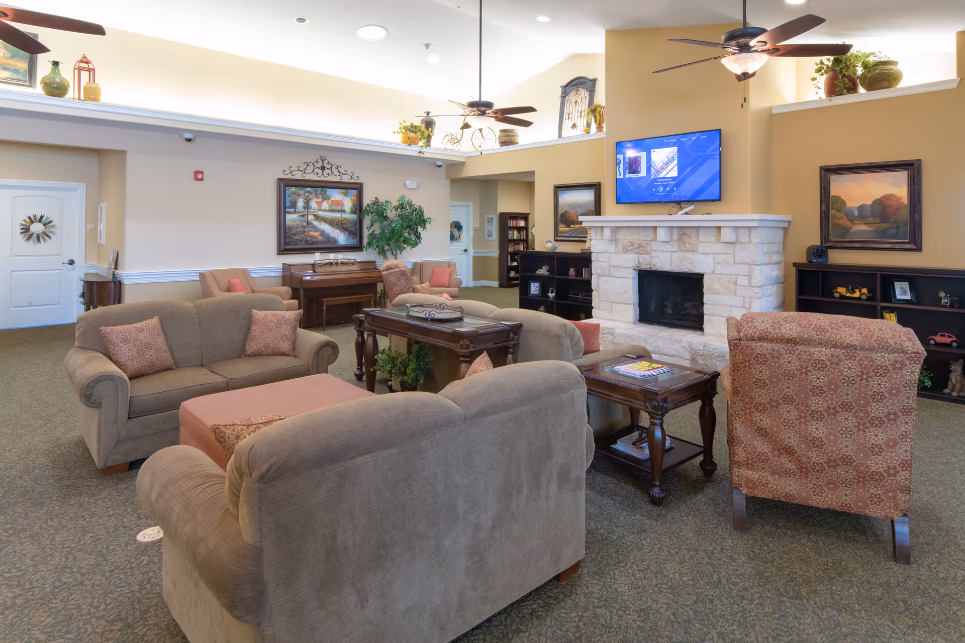 A cozy living room area in an assisted living facility featuring beige sofas and an armchair arranged around a wooden coffee table and side table. The room has a stone fireplace with a flat-screen TV mounted above it, ceiling fans with lights, framed landscape paintings on the walls, and decorative plants. There is a piano against the far wall and bookshelves with various items on display.