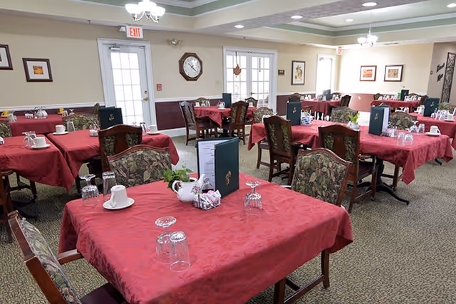 A dining room with multiple tables covered in red tablecloths, each set with cups, glasses, menus, and small flower arrangements. The room has beige walls, framed artwork, a clock, and two exit doors with windows. The ceiling features recessed lighting and chandeliers.