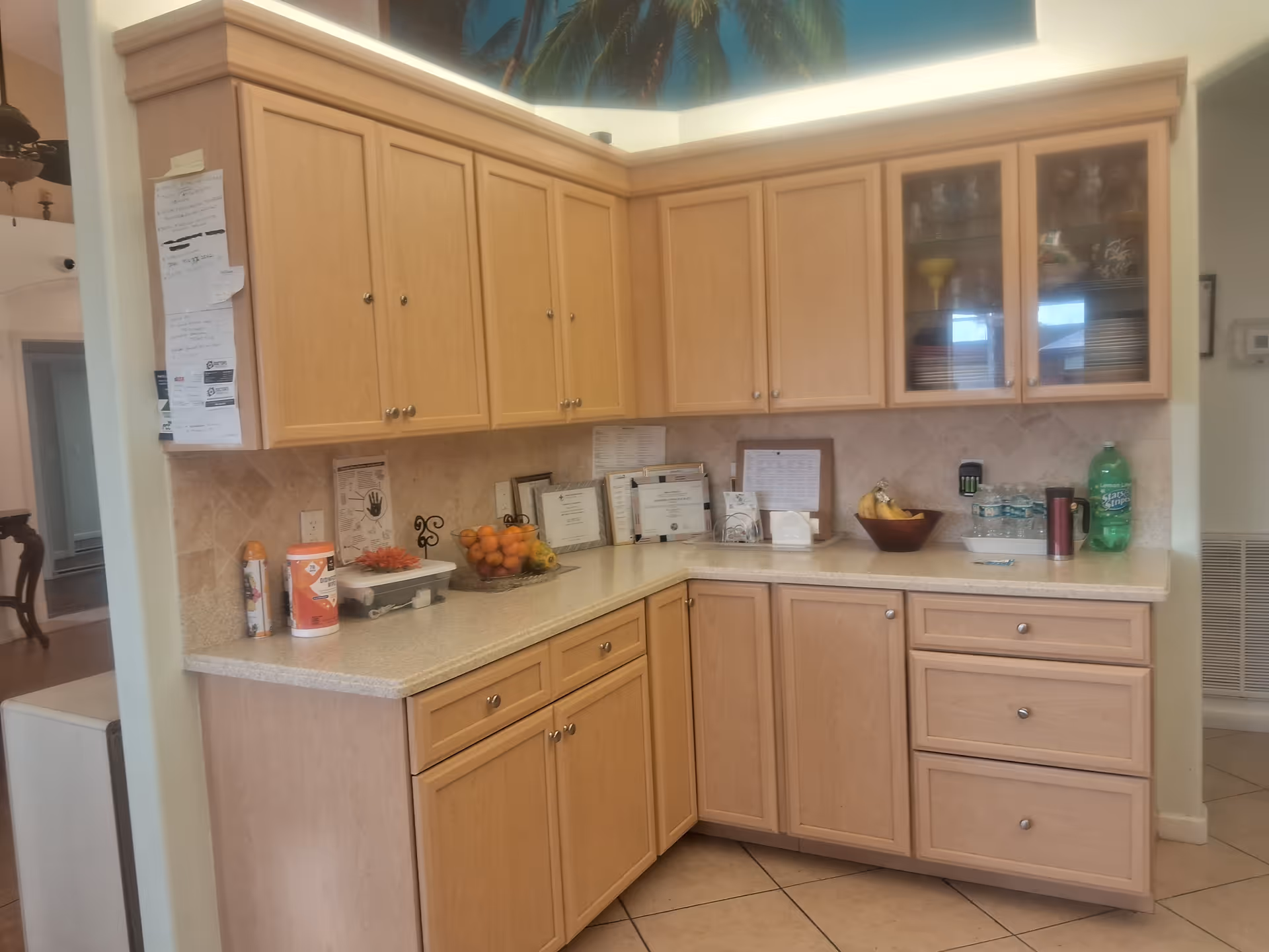 A kitchen corner with light wood cabinets and drawers, a beige countertop holding various items including a bowl of oranges, a bowl of bananas, bottled water, a green soda bottle, and some framed certificates or documents against the tiled backsplash.