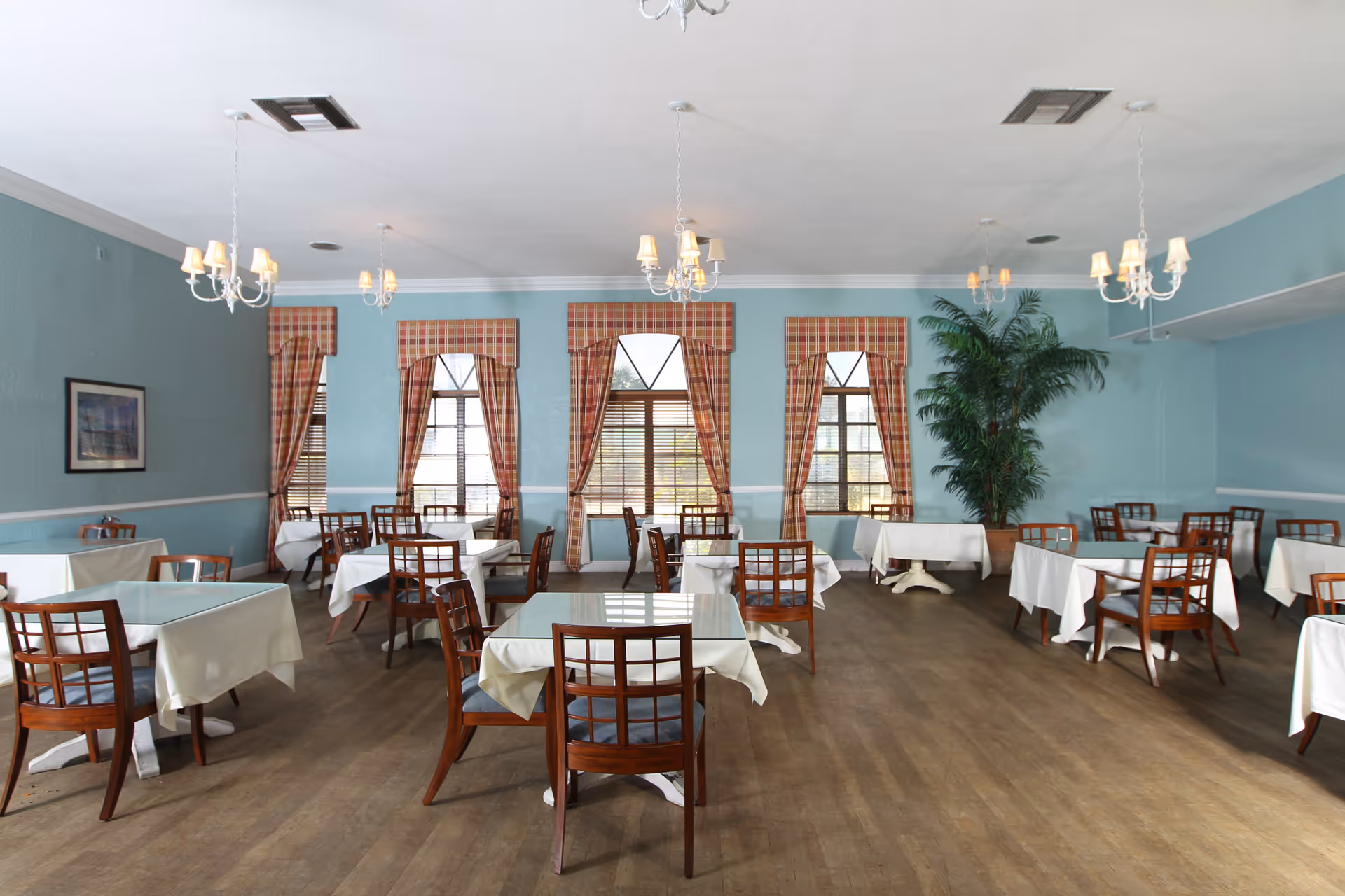 Spacious dining room with multiple tables covered in white tablecloths, wooden chairs, chandeliers, plaid window curtains, and a large potted plant.