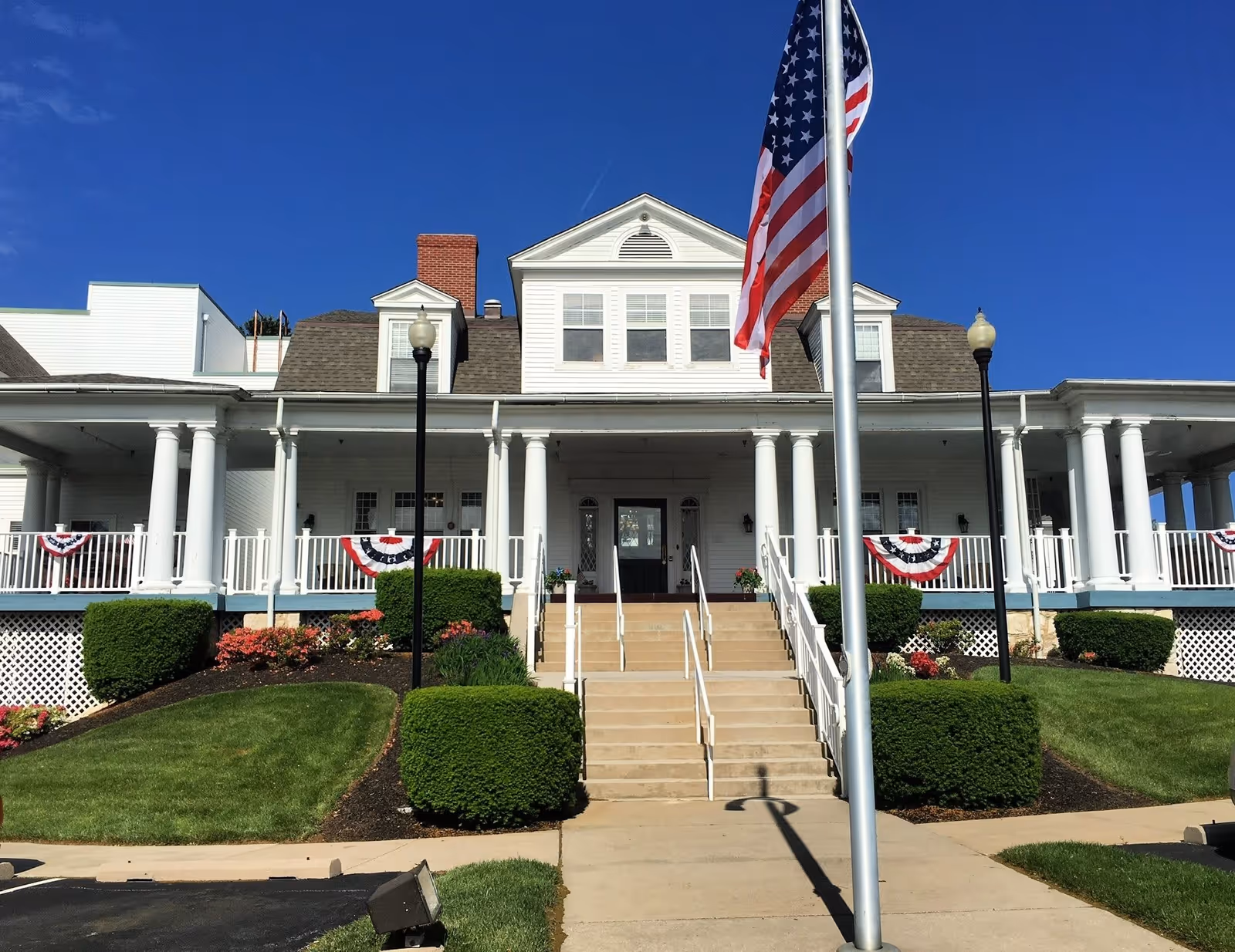 Front exterior view of a large white building with a wide porch supported by columns, decorated with patriotic red, white, and blue bunting. There are stairs leading up to the entrance, neatly trimmed bushes and flower beds on either side, and an American flag on a flagpole in the foreground against a clear blue sky.