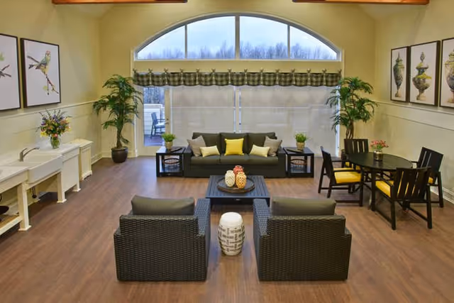 A spacious living room with wooden flooring, featuring a dark wicker sofa with yellow and beige cushions, two matching armchairs, a coffee table with decorative vases, and a small round dining table with four chairs. Large arched window with blinds and valance, potted plants, and framed artwork on the walls complete the room.
