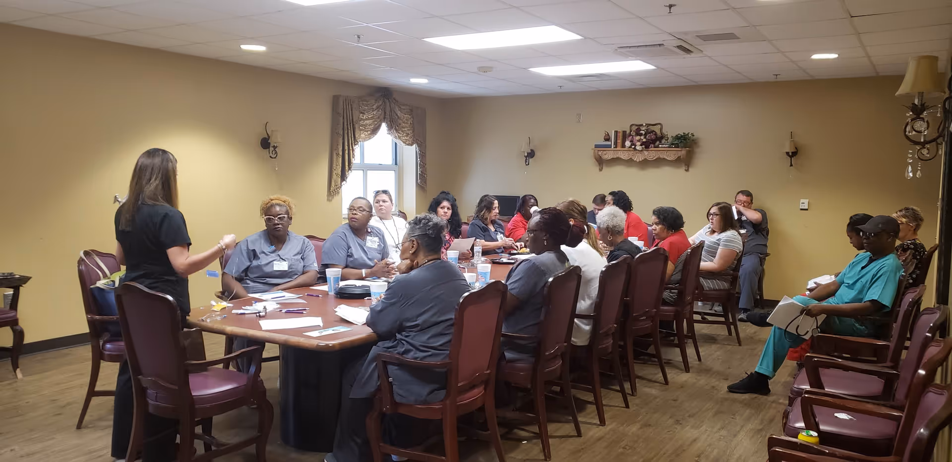 A group of healthcare workers seated around a large rectangular table in a meeting room. One woman stands at the head of the table speaking to the group. The room has beige walls, a window with curtains, wall sconces, and wooden flooring.