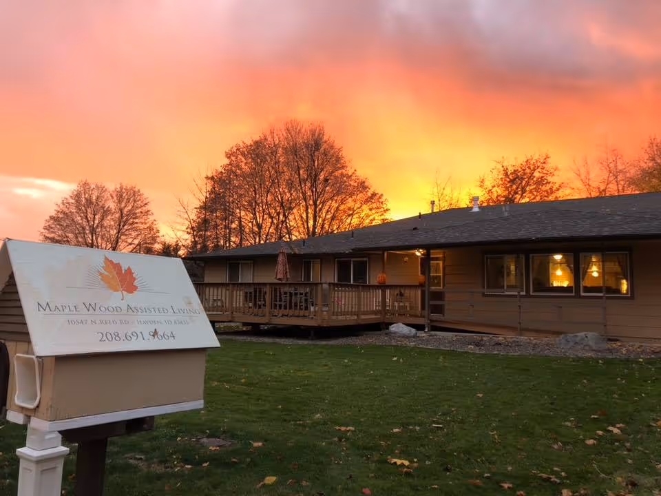 Exterior view of Maple Wood Assisted Living facility at sunset with a vibrant orange and pink sky. The building is a single-story structure with a wooden deck and several windows illuminated from inside. A sign in the foreground displays the facility's name, address, and phone number.