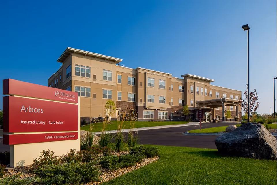 Exterior view of the Arbors assisted living and care suites facility, a three-story beige building with multiple windows, a covered entrance, and a landscaped lawn with rocks and plants. A red sign in the foreground displays the facility name and address.