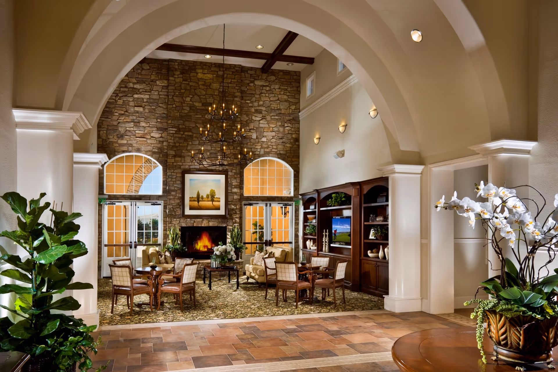 A spacious and elegant recreation center room with high arched ceilings and a stone fireplace at the far end. The room features comfortable seating with several armchairs and small tables arranged on a patterned carpet. There is a large wooden entertainment center with a television and decorative items. The room is well-lit with wall sconces and a chandelier hanging from the ceiling. Large windows with arched tops allow natural light to enter. Green plants and floral arrangements add a touch of nature to the space.