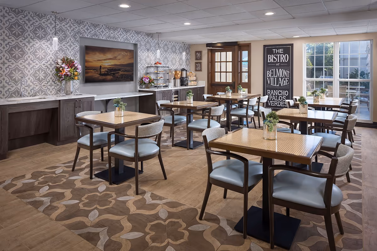 A bright and inviting dining room with several wooden tables and cushioned chairs arranged neatly. The room features patterned wallpaper, a decorative floor with floral designs, and a sideboard with flowers and a coffee station. Large windows allow natural light to fill the space, and a sign on the wall reads 'The Bistro at Belmont Village Rancho Palos Verdes.'