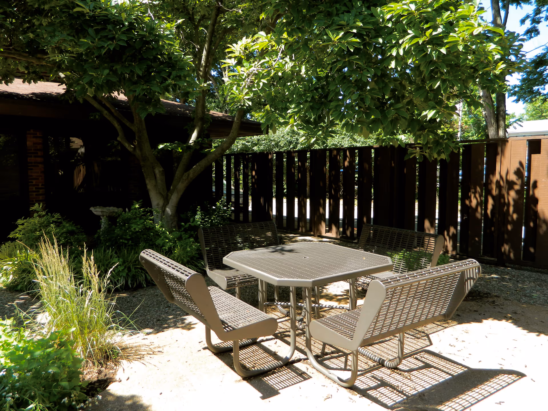 Outdoor patio area with a metal table and four attached benches under the shade of leafy trees, surrounded by greenery and a wooden fence.