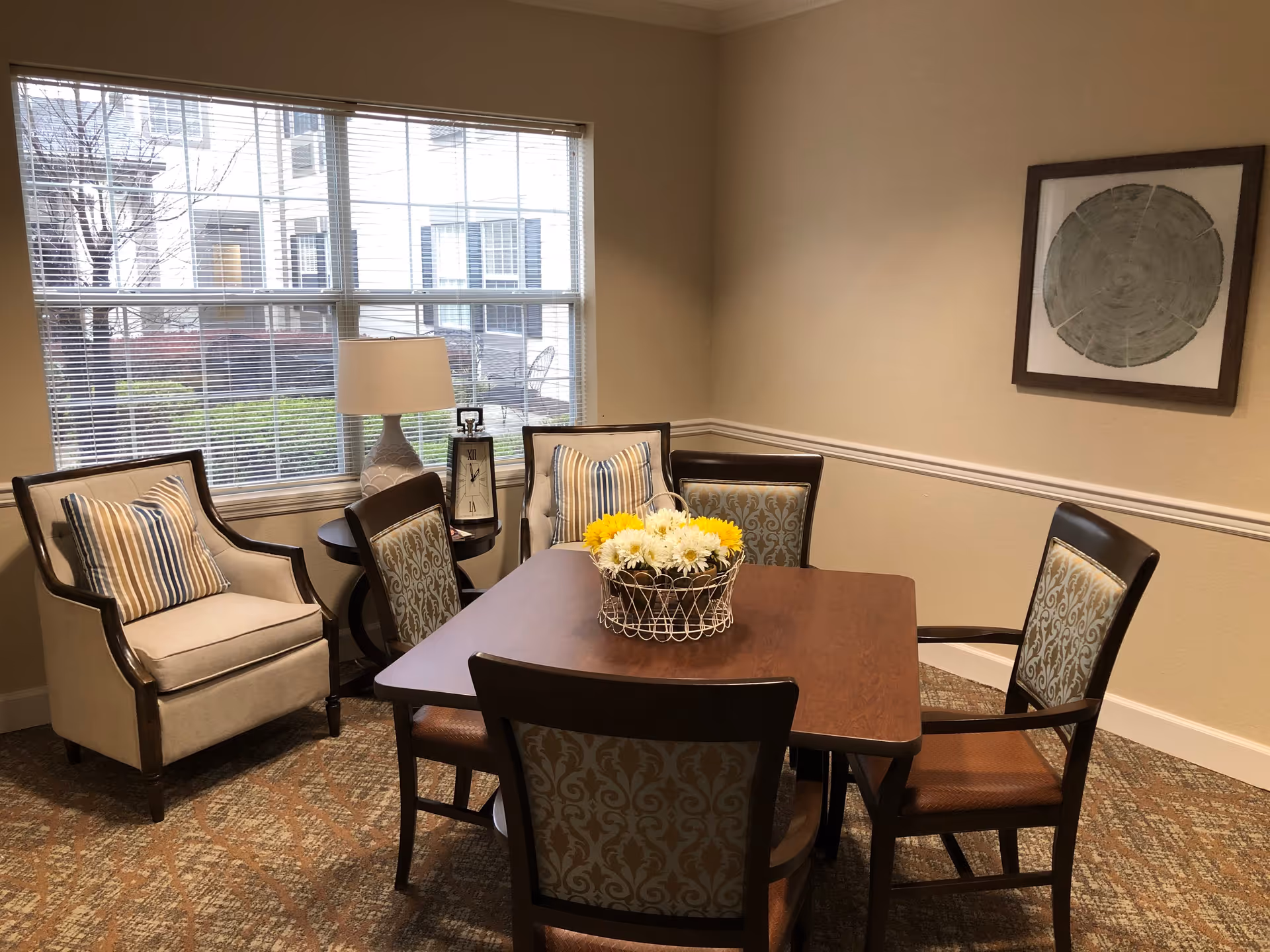 A cozy sitting area with a square wooden table surrounded by four upholstered chairs with patterned backs. Two cushioned armchairs with striped pillows are placed near a window with blinds. A small round side table holds a lamp and a decorative clock. A framed artwork hangs on the beige wall, and a basket of yellow and white flowers sits on the table. The room has patterned carpet flooring.