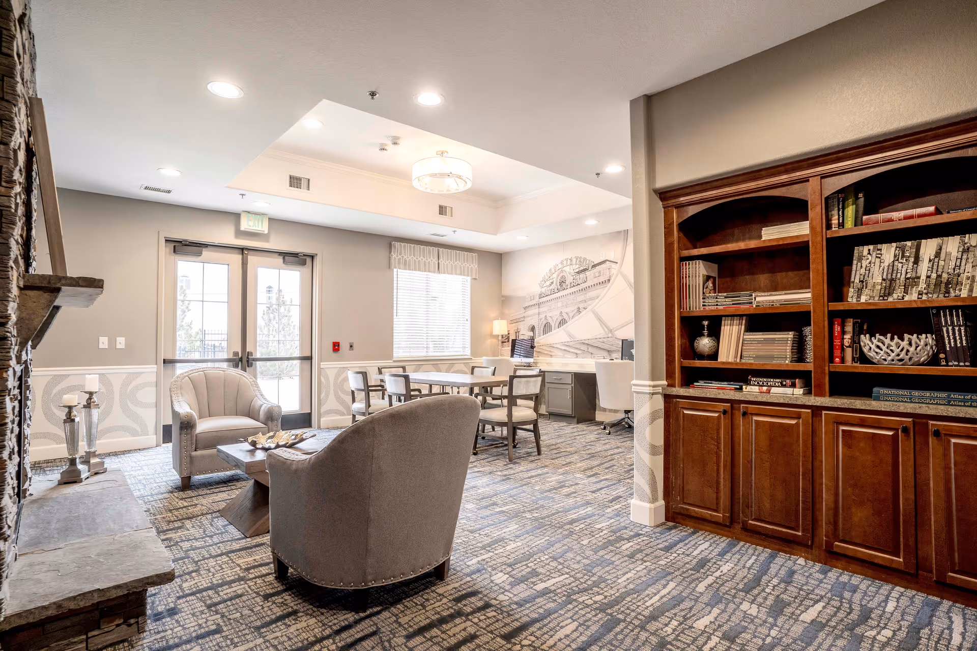 A bright and spacious living room area in an assisted living facility featuring two upholstered armchairs around a wooden coffee table, a stone fireplace on the left, a large wooden bookshelf filled with books on the right, and a table with chairs near a window in the background. The room has patterned carpet flooring, neutral-colored walls, and recessed lighting in the ceiling.