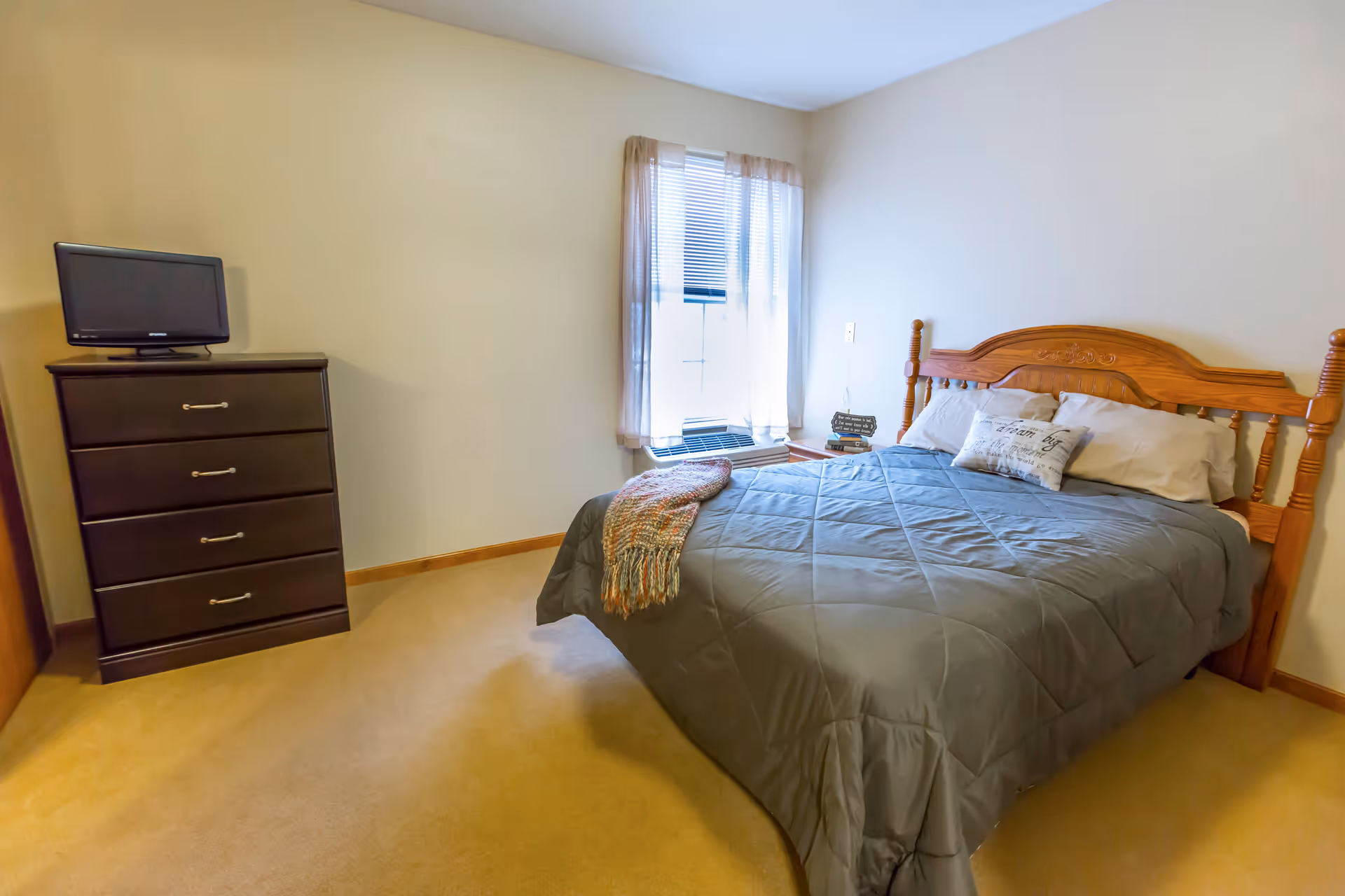 A simple bedroom with a wooden bed frame, a gray comforter, and pillows. There is a window with sheer curtains letting in natural light. Next to the bed is a small nightstand with books and a decorative sign. Across from the bed is a dark wooden dresser with a small flat-screen TV on top. The room has beige carpet and light-colored walls.