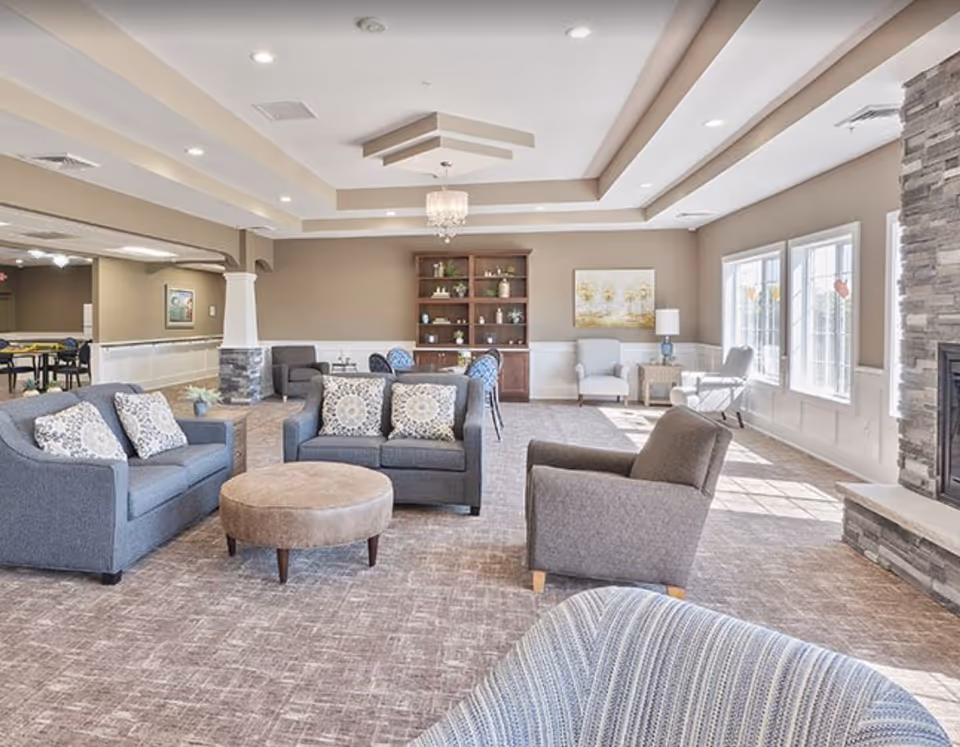 A spacious and well-lit living room area in an assisted living facility featuring multiple comfortable armchairs and sofas with patterned cushions, a round ottoman in the center, a stone fireplace on the right, large windows allowing natural light, a wooden bookshelf with decorative items, and a chandelier hanging from a tray ceiling.