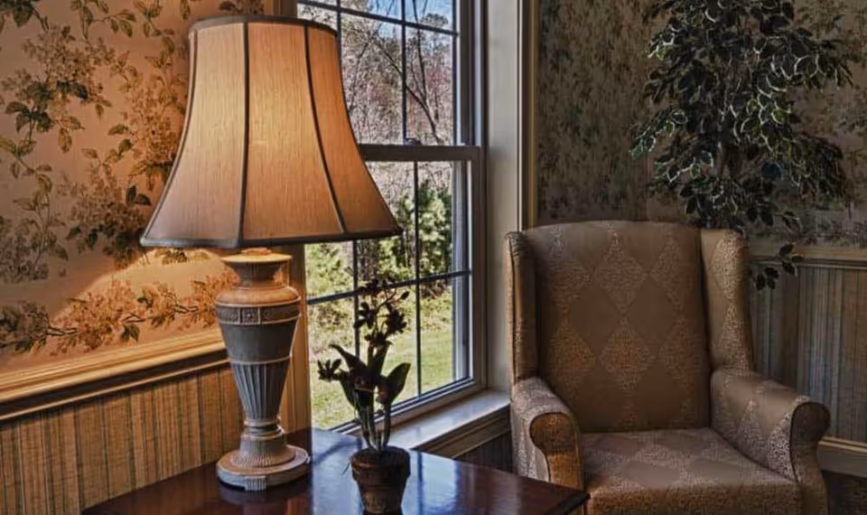 A cozy corner of a room featuring a patterned armchair next to a window with a view of greenery outside. A table beside the chair holds a decorative lamp with a beige shade and a small potted plant. The walls are adorned with floral wallpaper and wood paneling.
