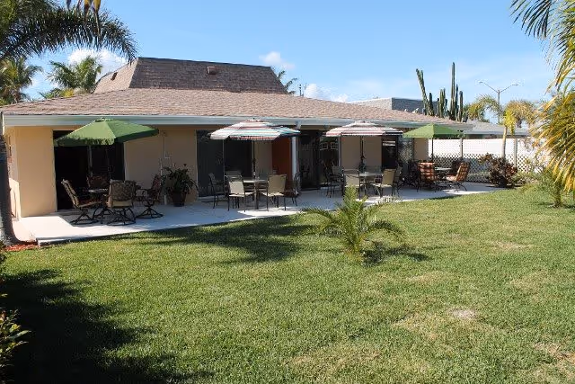 Outdoor patio area of a single-story building with several tables and chairs under green and striped umbrellas, surrounded by palm trees and a well-maintained grassy lawn under a clear blue sky.
