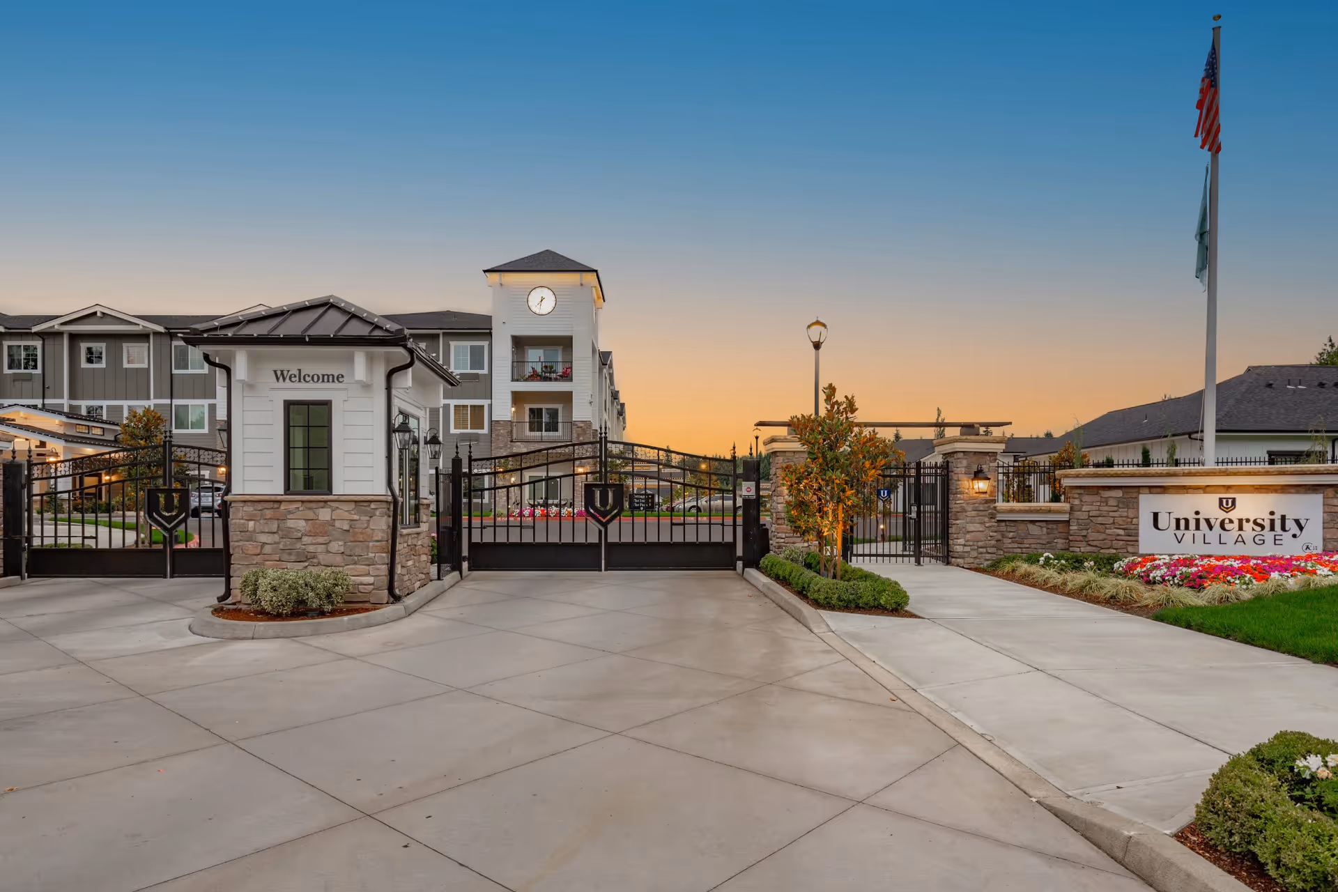 Gated entrance with a small welcome guardhouse and a sign reading "University Village" in front of a multi-story senior living building at sunset.