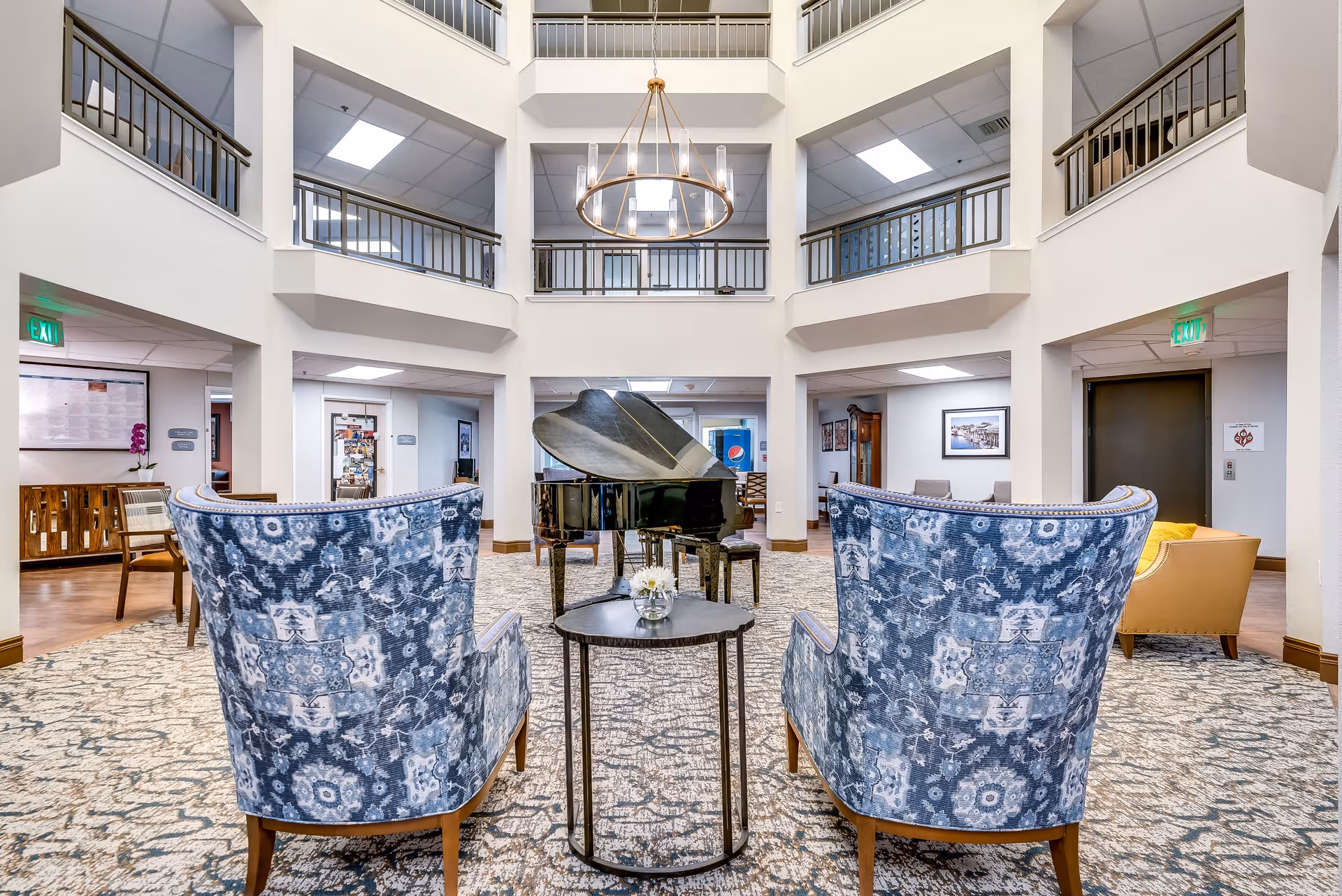A spacious and well-lit senior living facility common area with two blue patterned armchairs facing a black grand piano. The room features a high ceiling with a modern chandelier, a patterned carpet, and a second-floor balcony overlooking the area. There are additional seating areas and doors leading to other rooms.