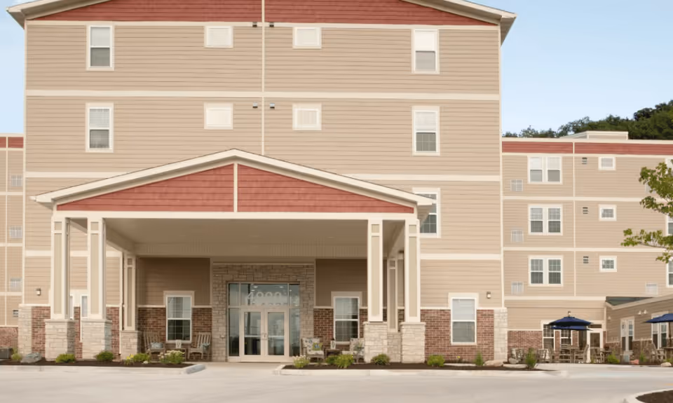 Exterior front view of a multi-story senior living facility building named Serenity, featuring a covered entrance with stone and brick accents, multiple windows, and outdoor seating areas with umbrellas on the right side.