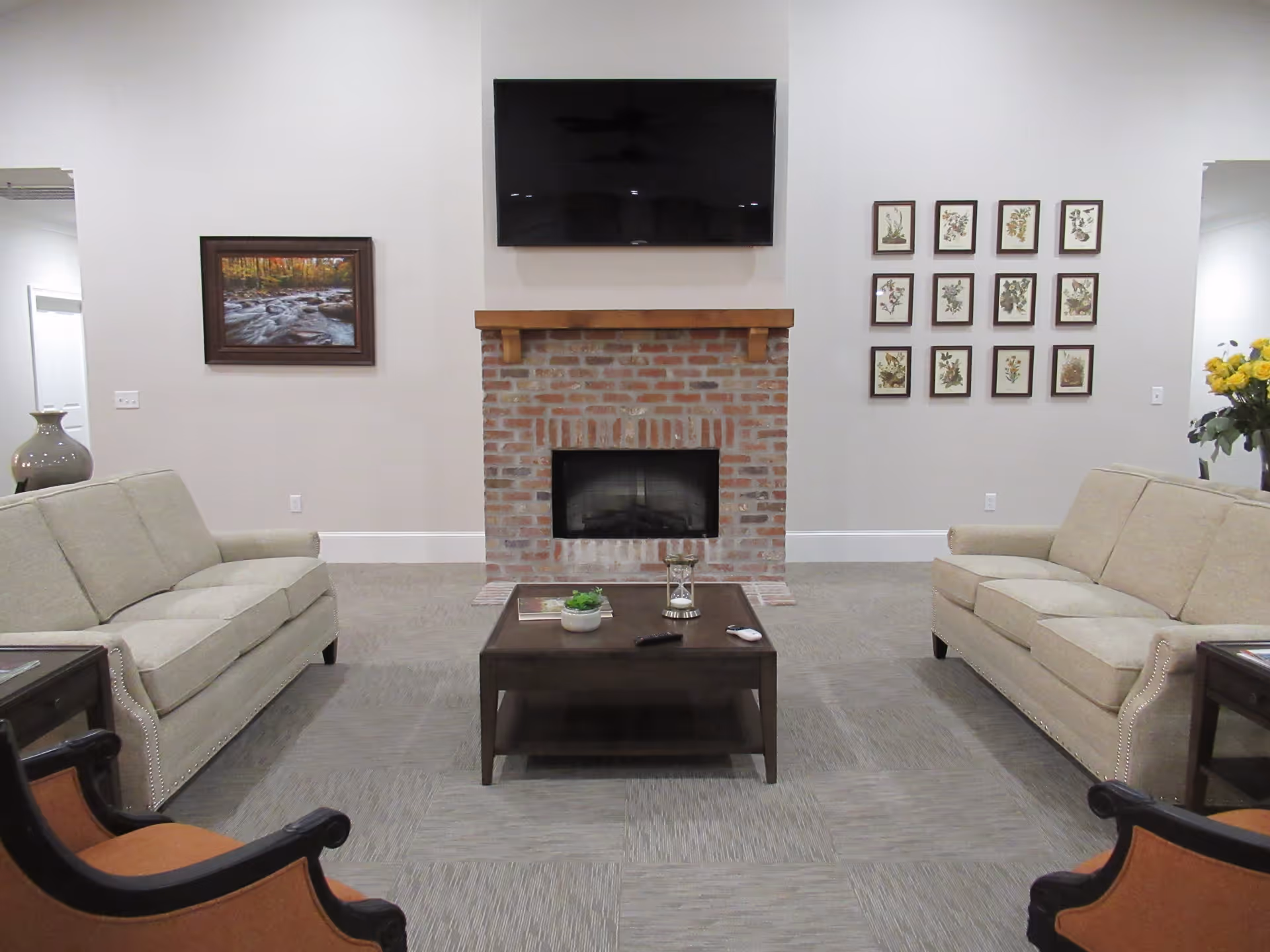 A cozy living room with two beige sofas facing each other, a dark wooden coffee table in the center, and a brick fireplace with a wooden mantel. Above the fireplace is a mounted flat-screen TV. The walls are decorated with framed pictures, including a landscape painting on the left and a grid of botanical prints on the right. There are also two armchairs with orange cushions partially visible in the foreground.