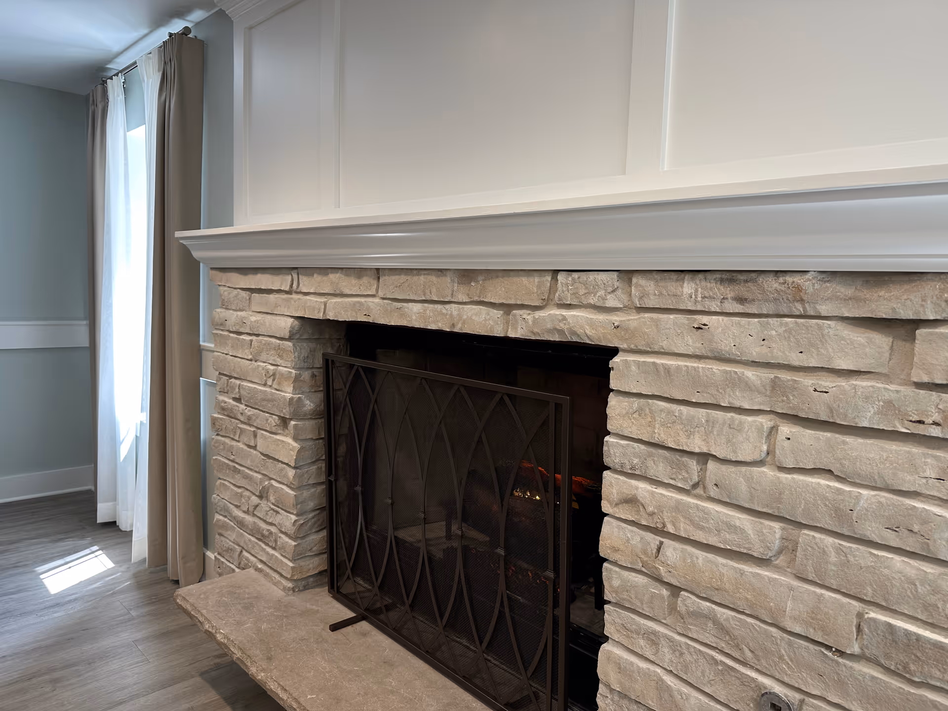 Stone fireplace with a black metal screen in a room with light-colored walls and wood flooring. A window with sheer white curtains and beige drapes is visible to the left.