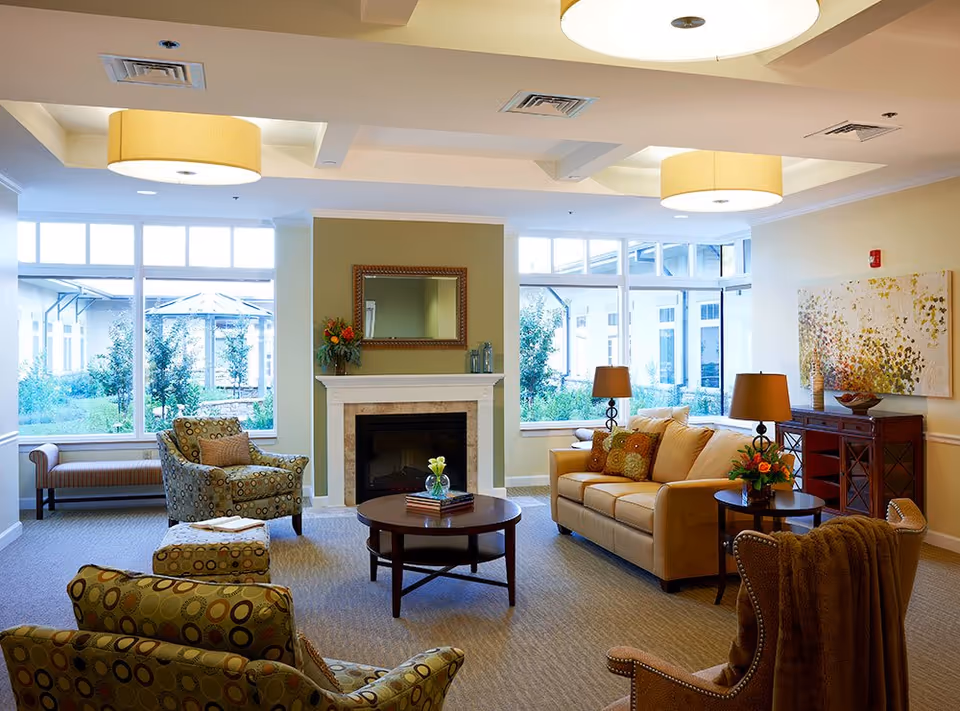 A cozy living room area with large windows letting in natural light. The room features a beige sofa with decorative pillows, two patterned armchairs, a round wooden coffee table with a small flower vase and books, a fireplace with a mirror above it, and a wooden sideboard with a lamp and artwork on the wall. The room has soft lighting from ceiling fixtures and table lamps, with a view of greenery outside the windows.