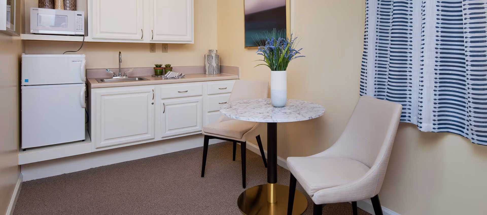 Small kitchenette with white cabinets, a mini fridge and microwave, and a round marble-top table with two upholstered chairs.